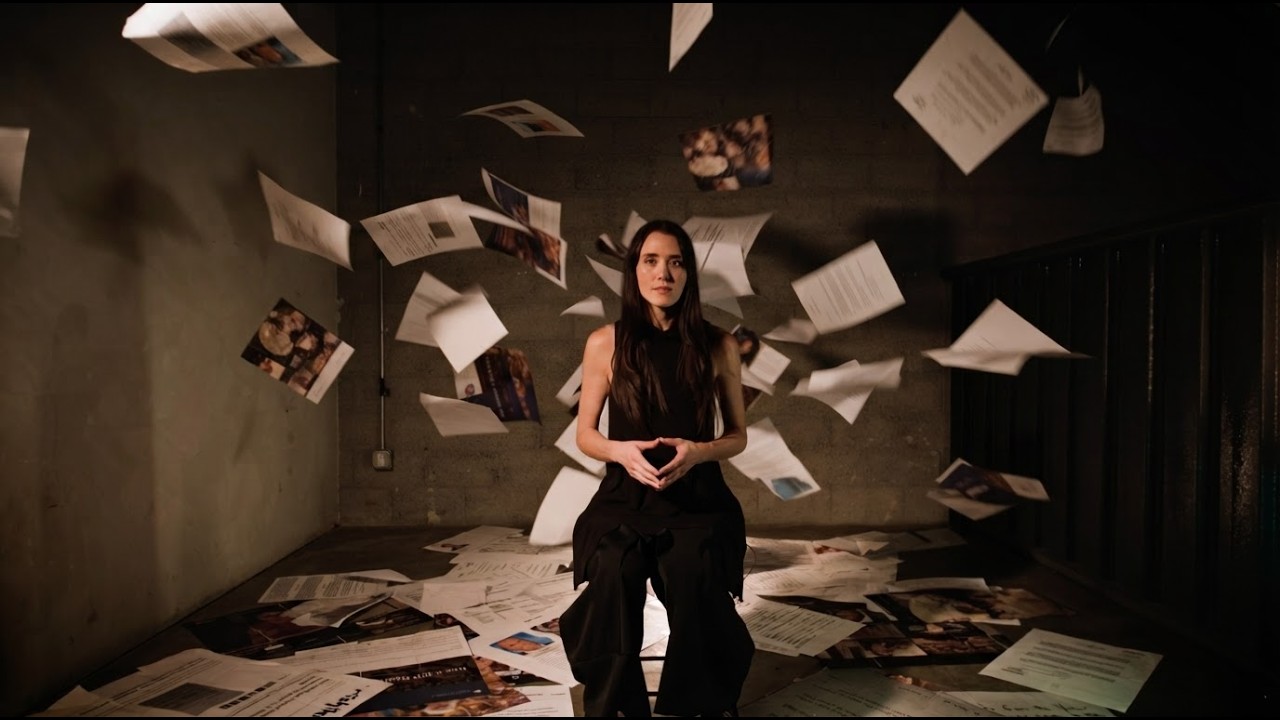 Myra Nishizaki seated as printed pages fall around her, the documentary&rsquo;s title frame